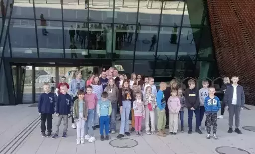 Grupa dzieci i opiekunów pozuje do wspólnego zdjęcia przed nowoczesnym budynkiem z przeszkloną fasadą